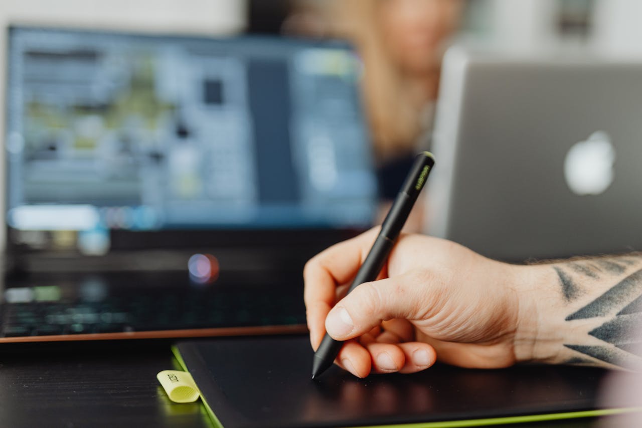 Close-up of a person using a stylus pen on a digital pad with a blurred laptop background.